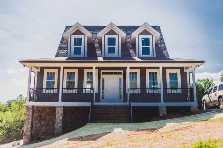 Cape cod style home with three dormers over a hip roof front porch. The house is a gray blue tone with white windows and trim.