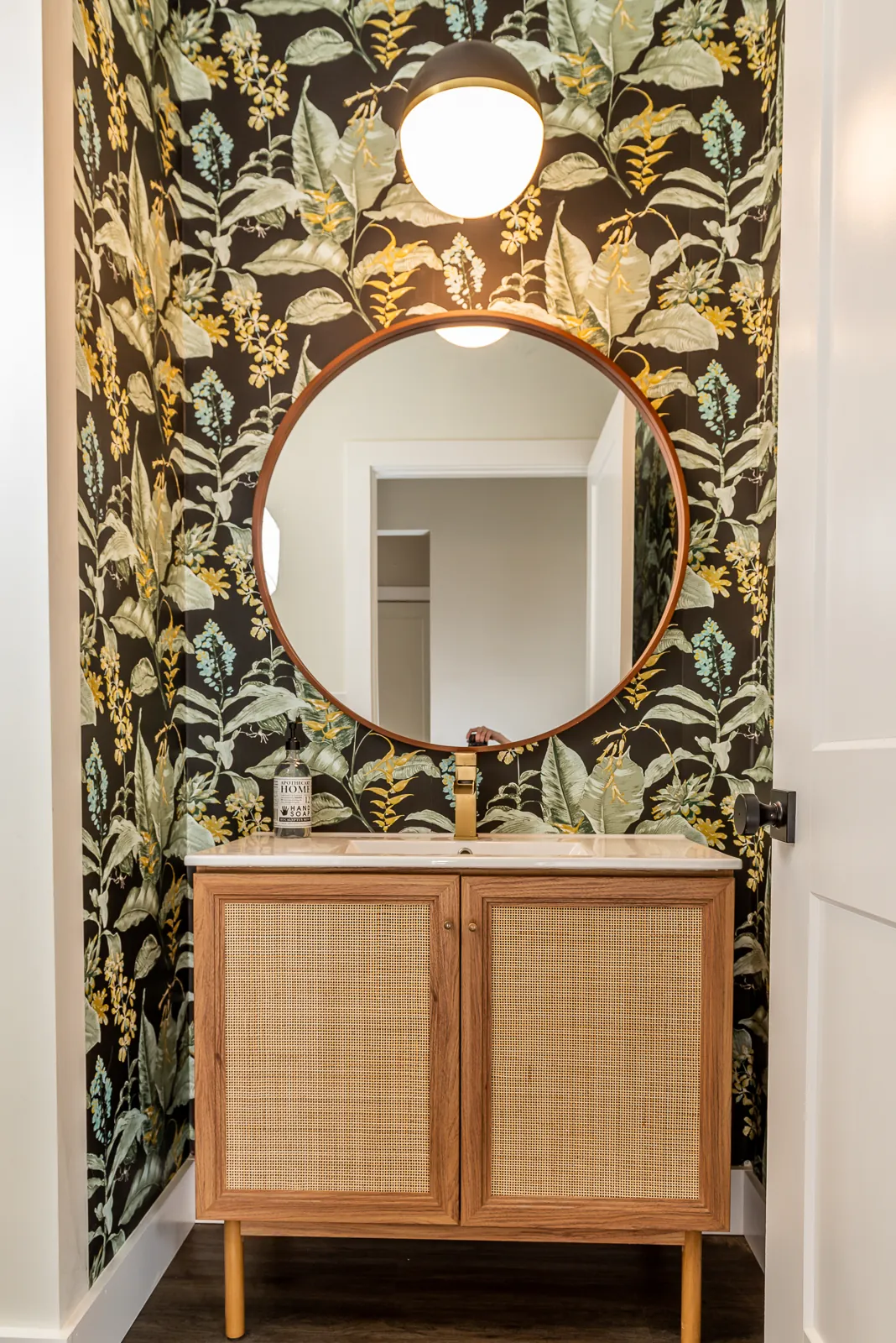 Powder room with botanical wallpaper, round mirror and rattan vanity in the LynBrook Home