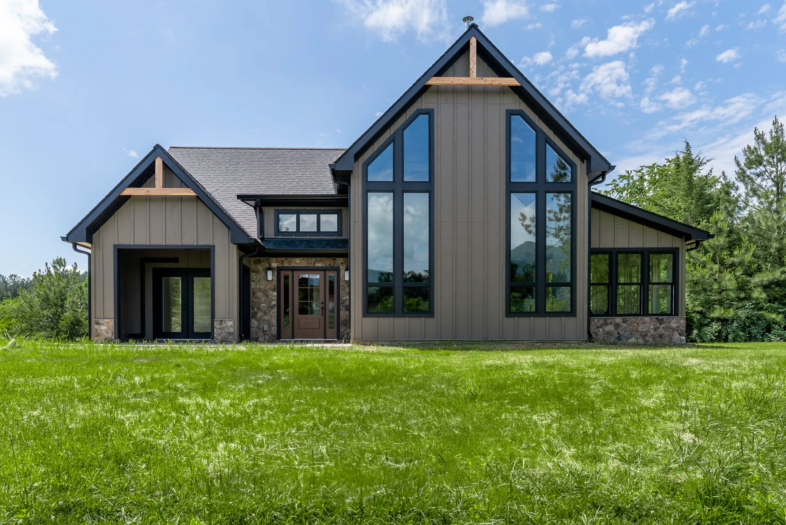 Front exterior view of a beautiful taupe A frame style home with large windows, black trim and gutters. Accents of natural stone surround the door and the lower portion of the foundation and wood accents are in the gables.