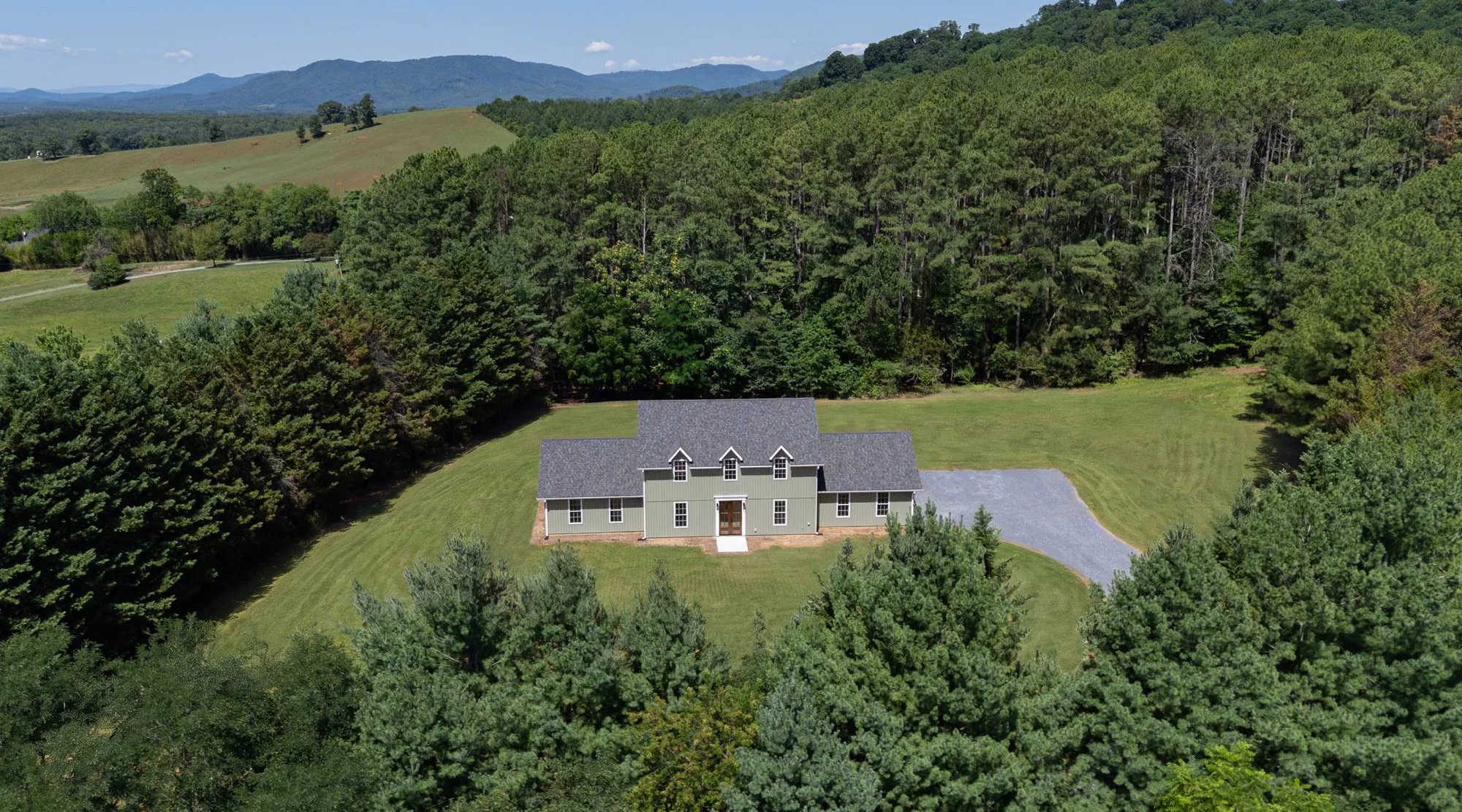 Aerial view of a sage green farmhouse with three dormers in a clearing of pine woods, with the Blue Ridge Mountains beyond, near Bedford, VA